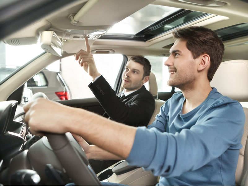 Two men are sitting in a car, with one pointing at the dashboard controls.