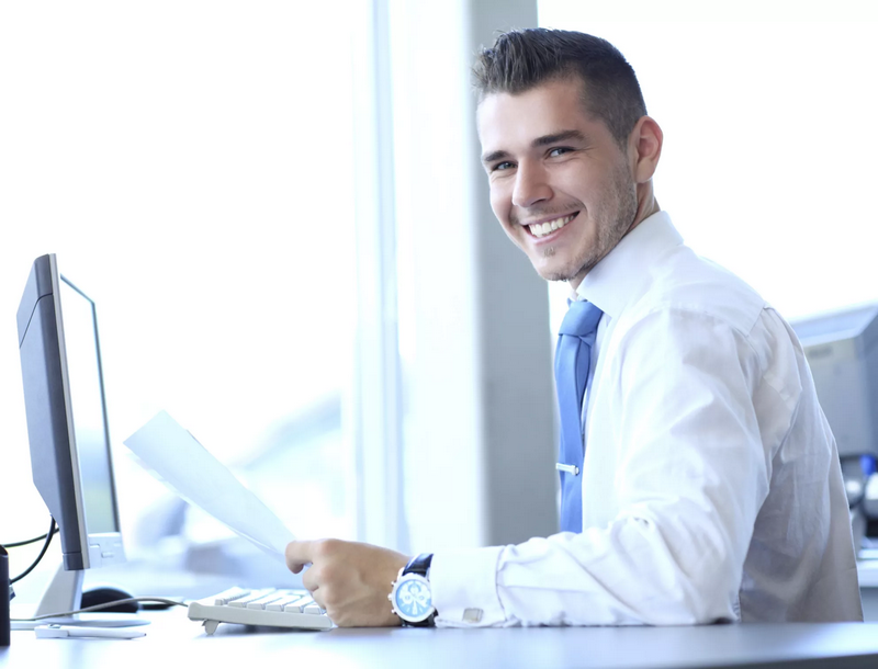 A man in a white shirt and blue tie is sitting at a desk with a computer, holding papers, and smiling.