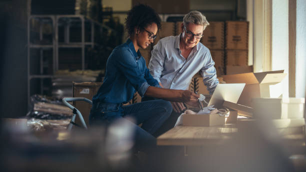 Two small business owners collaborate while reviewing inventory and using a laptop in their workspace.