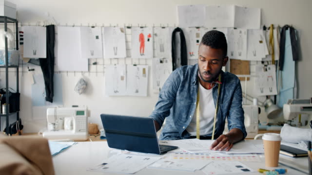 Small business owner reviewing documents and working on a laptop in a busy office with design sketches on the wall.