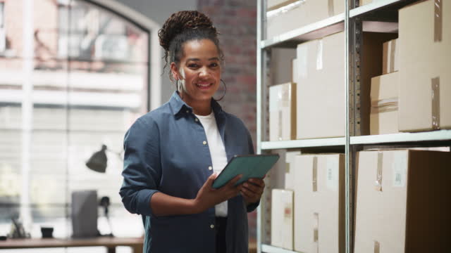 Small business owner in warehouse with packing boxes, holding a tablet and smiling, ready to recover.